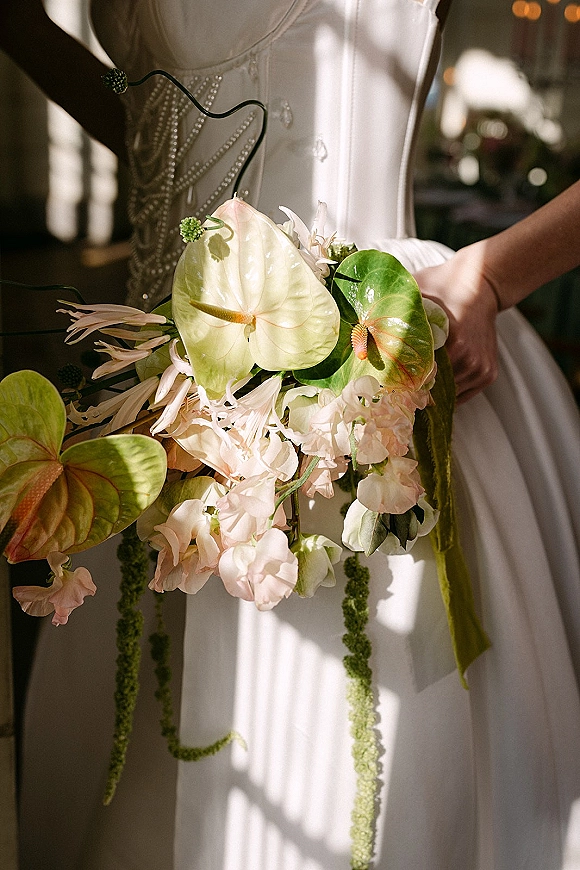 Bridal bouquet with tropical bridal bouquet styling, green anthurium and blush blooms, long ribbon tails against a beaded bodice in warm indoor light