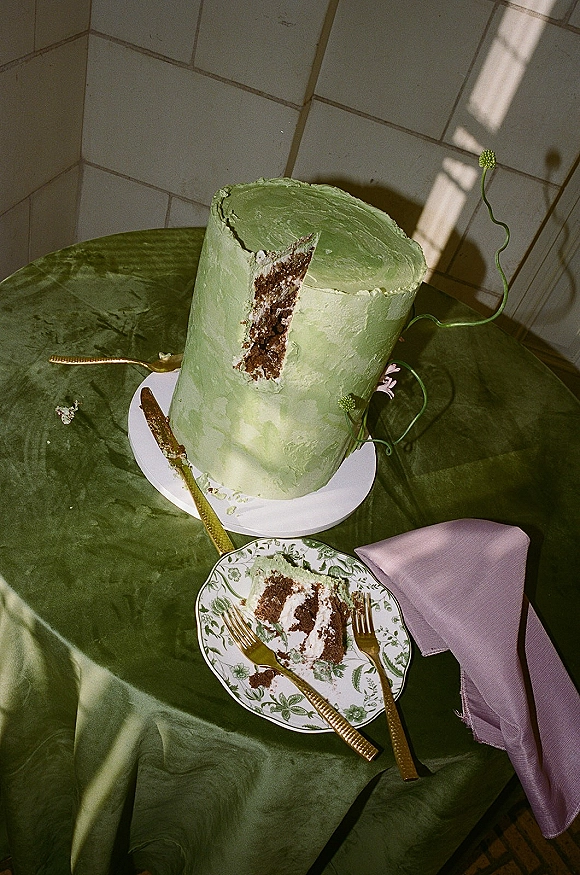 Wedding cake with green wedding cake frosting, a slice on a floral plate beside a gold cake knife, on a green tablecloth in window light shadows