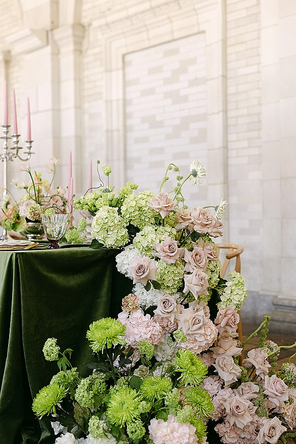 Reception tablescape with wedding floral tablescape on a green velvet tablecloth, pink taper candles, silver candelabra, and glass goblets by a stone wall