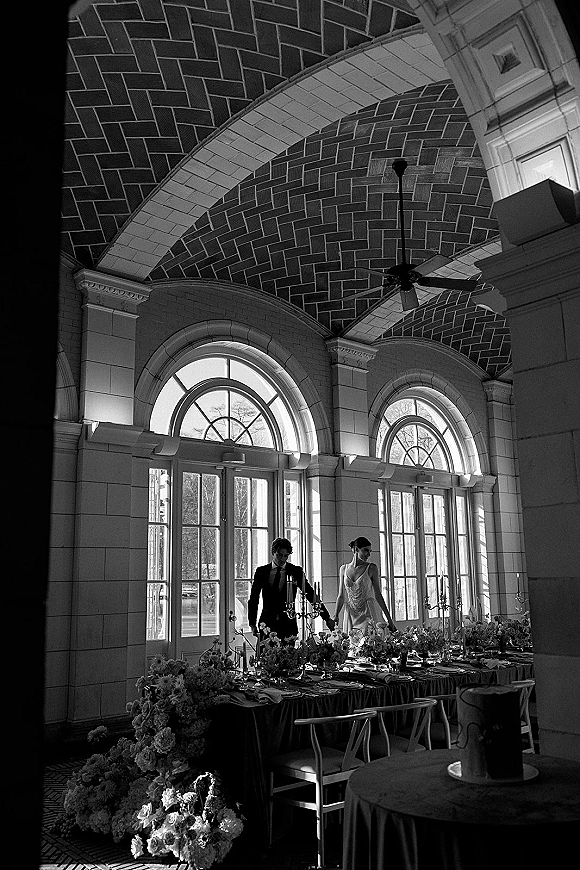 Reception tablescape with long banquet table, floral garland and taper candles, set under arched windows in a stone-walled venue.