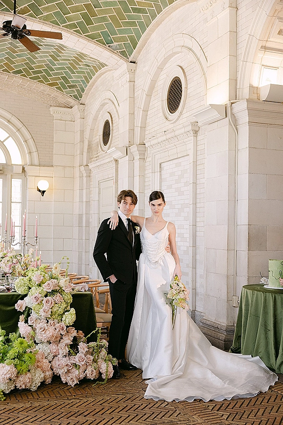 Couple portrait of bride holding bouquet beside groom in black tuxedo, set by a reception table under an arched brick wall backdrop.