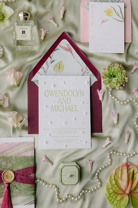 Wedding invitation suite flat lay with illustrated cards, burgundy envelopes, pearls, ring box and perfume on sage green fabric backdrop