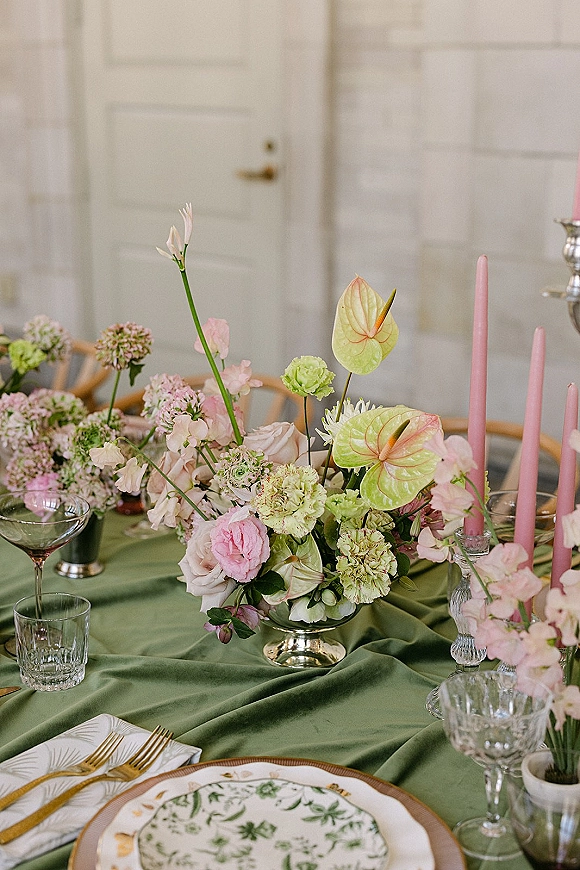 Reception tablescape with green wedding tablecloth, pastel floral centerpiece, pink taper candles, crystal goblets, and gold flatware by a white paneled wall