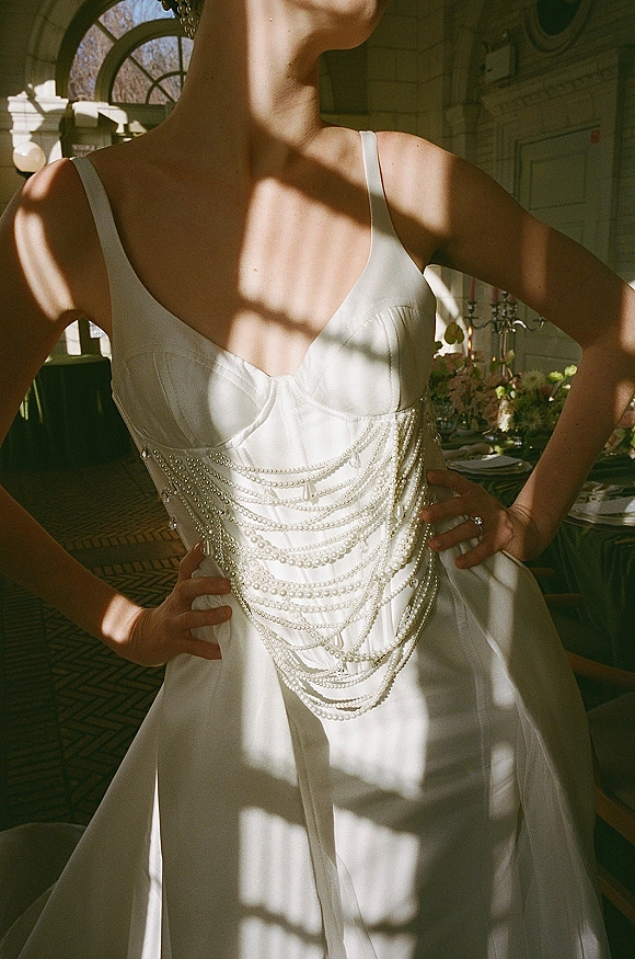 Bridal gown detail with pearl wedding dress draped bodice and satin spaghetti straps, hands on hips by arched windows casting shadows