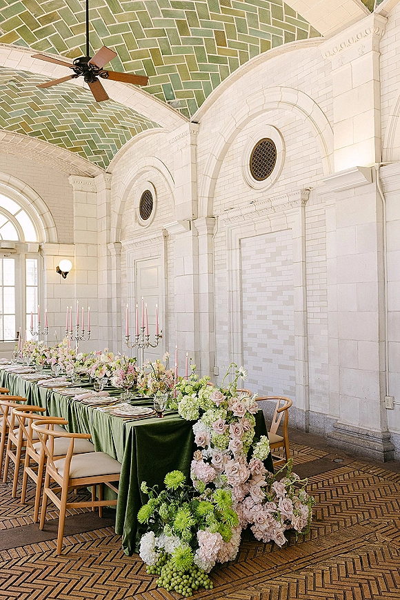 Reception tablescape with long banquet table decor on green velvet tablecloth, pastel flowers, silver candelabras, pink tapers in arched brick hall