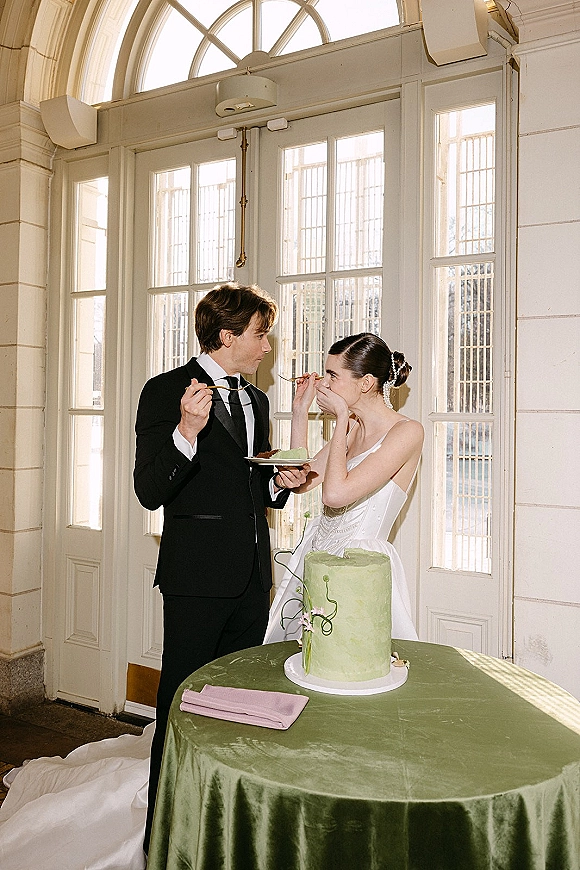 Wedding cake cutting as bride in white gown and groom in black tuxedo share a green cake at a round table by arched windows.