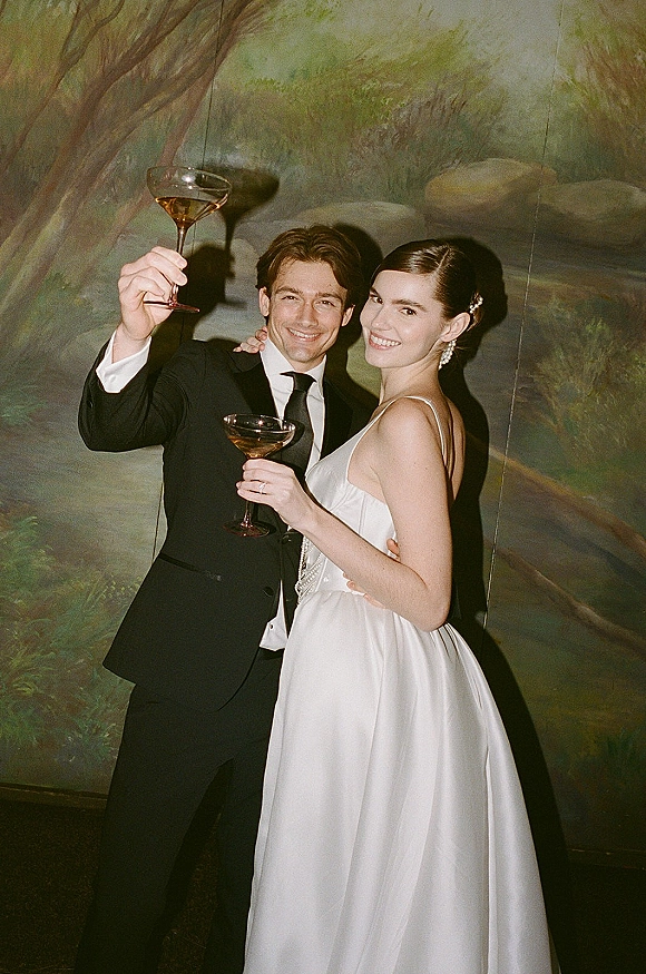 Wedding couple portrait of newlyweds raising champagne coupes in front of a painted mural backdrop, bride in satin gown and groom in black suit