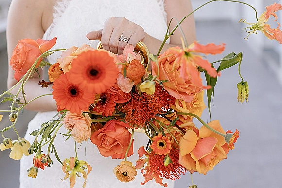 Bridal bouquet with orange wedding bouquet blooms in peach roses and ranunculus, ribbon-wrapped with wedding rings against a neutral wall