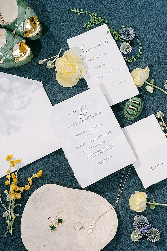 Invitation suite wedding invitation flat lay with deckled-edge cards, envelopes, rings, emerald earrings and green heels on blue fabric backdrop