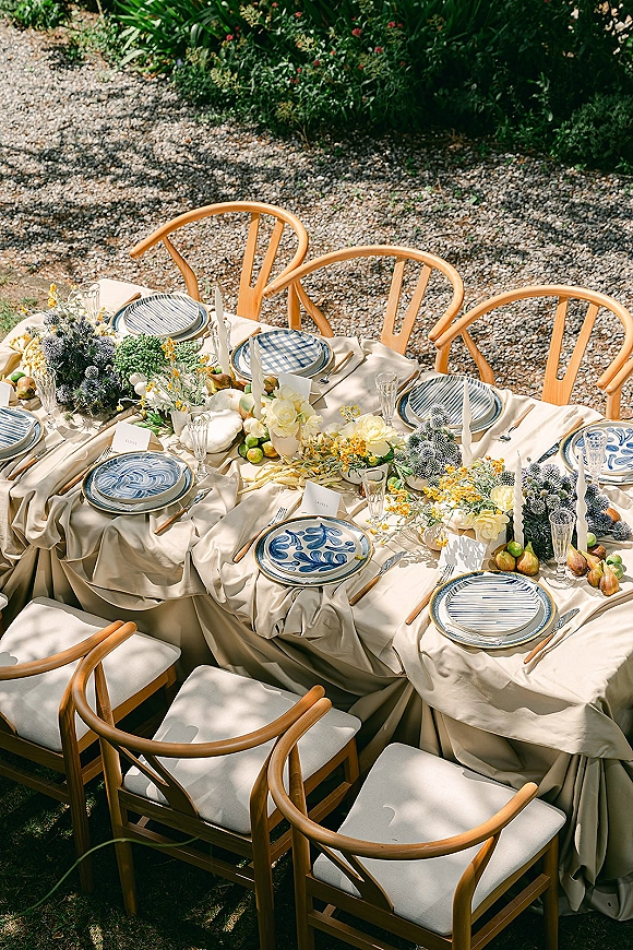 Reception tablescape with an outdoor wedding table dressed in neutral linens, patterned plates, place cards, glassware, florals, pears and limes amid garden greenery