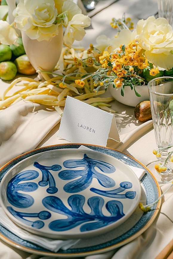Reception tablescape with wedding place card, blue patterned plates, linen napkin, glass goblet, and white rose centerpiece with yellow blooms
