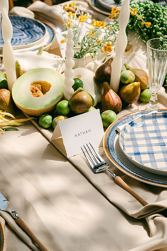 Reception tablescape with wedding place setting of blue patterned plates, place card, taper candles, and a fig and melon centerpiece in natural light