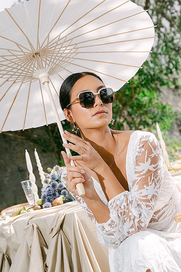 Bridal portrait of a bride with parasol wearing sunglasses and a white lace gown, seated by a styled table in garden greenery and stone wall backdrop