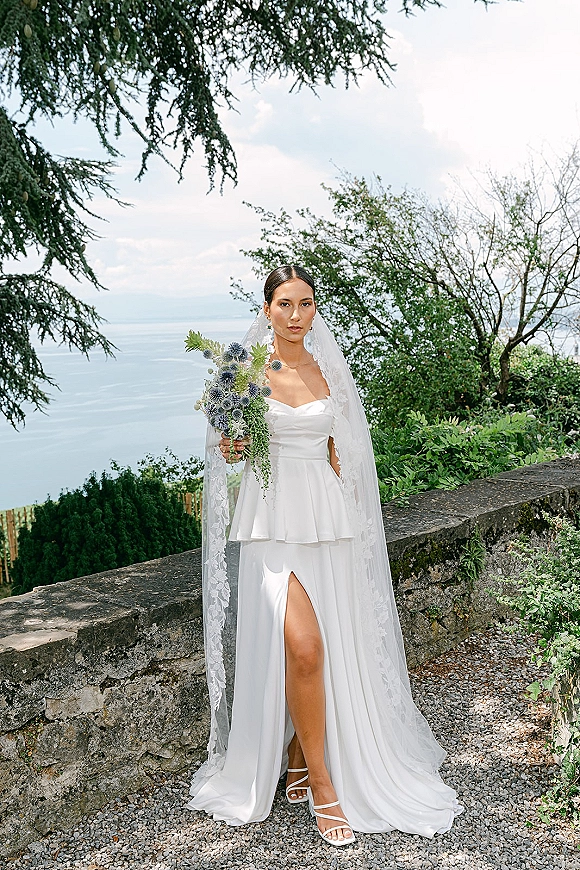 Bridal portrait of a bride holding bouquet with a lace-edge veil and strapless dress, standing by a stone wall near a lakeside path
