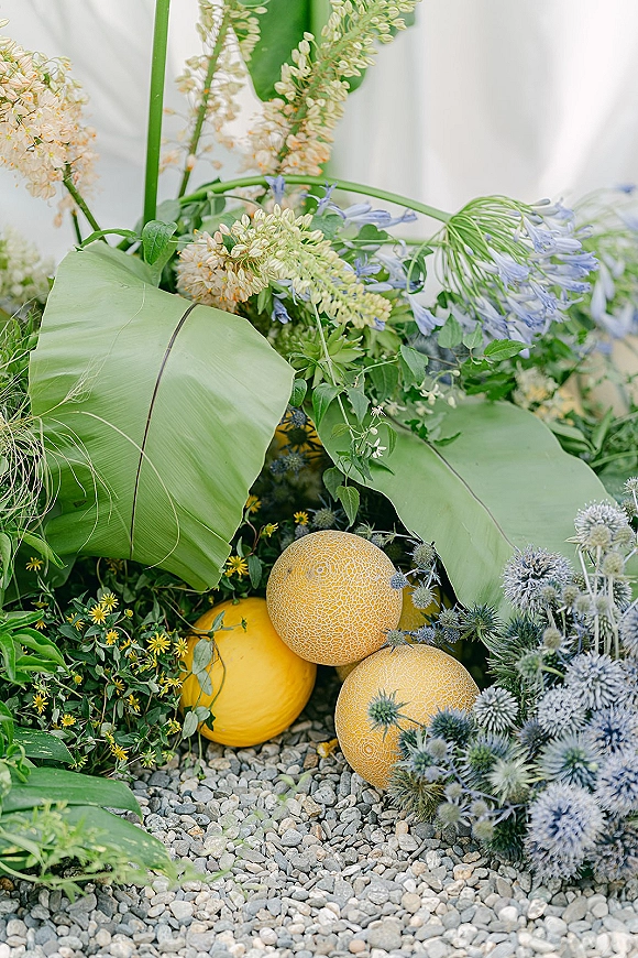 Floral arrangement with tropical leaf centerpiece featuring melons, blue thistle, and purple blooms on gravel before a white drape backdrop