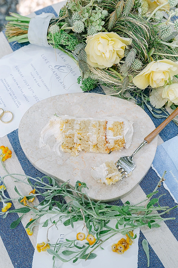 Wedding cake slice with buttercream frosting on a plate and fork beside a wedding invitation, yellow roses, and ring on blue striped linen