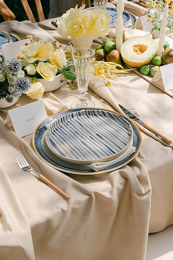 Reception tablescape with wedding place setting featuring striped and blue plates, beige linens, taper candles, and yellow rose centerpiece with fruit display outdoors