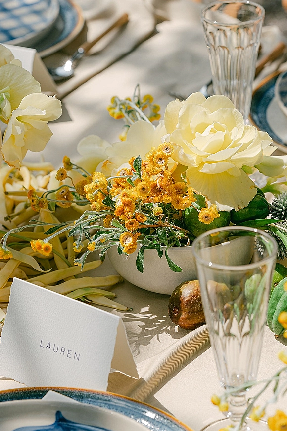 Reception tablescape with a wedding table centerpiece of yellow florals in a low white bowl, blue-rim plates, place card, and pear in sunlit shadows