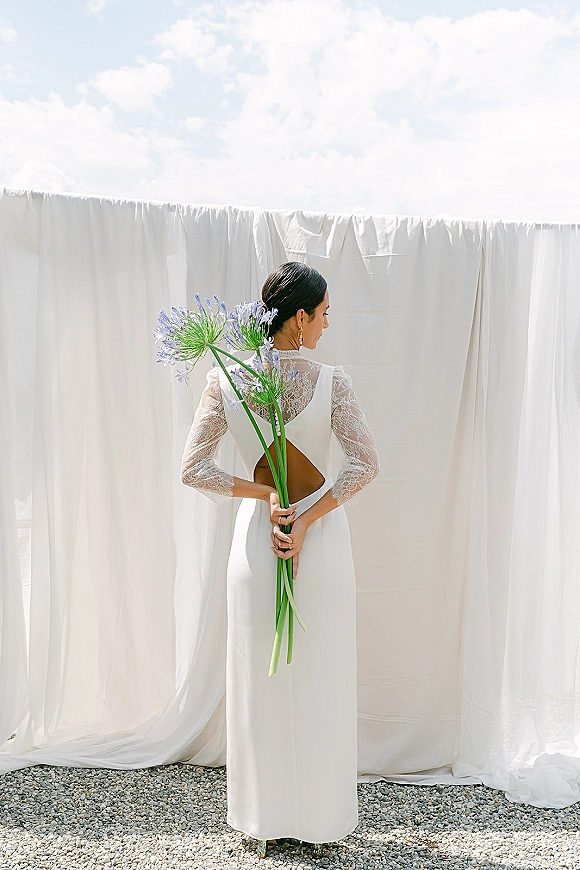 Bridal portrait of a bride holding bouquet behind back in a backless long sleeve lace wedding dress, against a white fabric backdrop and sky