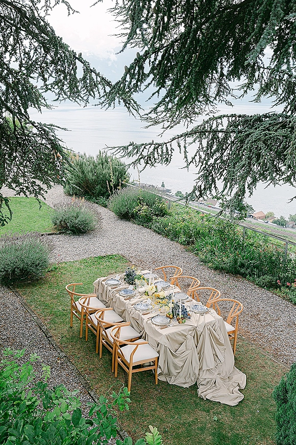 Reception tablescape with an outdoor wedding reception table set on beige linens, wooden chairs, floral centerpiece, taper candles, grapes, and lake view backdrop