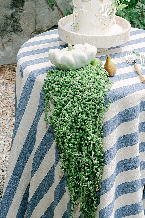 Wedding cake with floral piped buttercream on a stone stand, styled with trailing greenery, pears and a white pumpkin by a stone wall