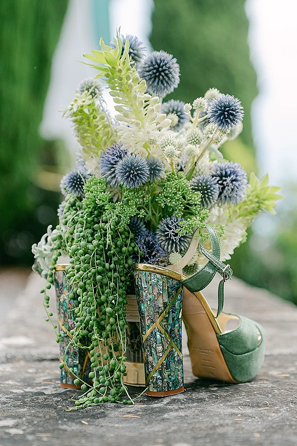 Bridal shoes in green wedding heels with patterned block heels beside blue thistle, succulents, and ribbon on stone pavement outdoors