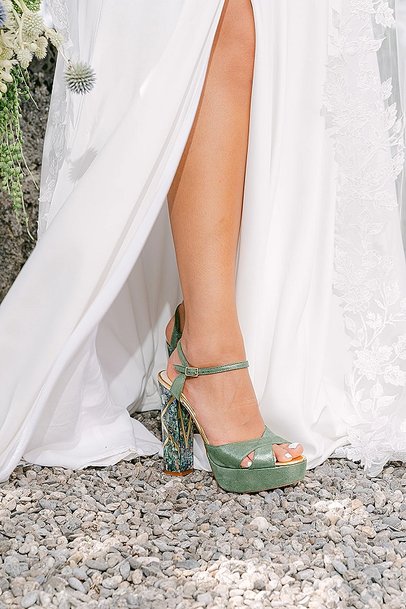 Bridal shoes in green wedding heels with ankle straps and patterned block heels beneath a lace dress hem on gravel by greenery