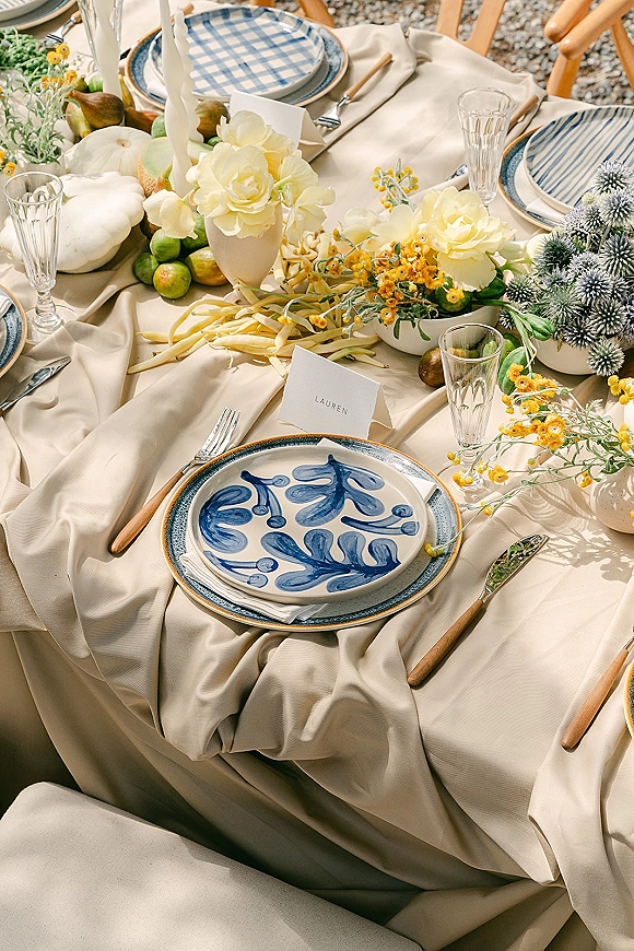 Reception tablescape with wedding place setting, blue patterned plates, taper candles, fruit and floral centerpieces on linen outdoors on gravel ground