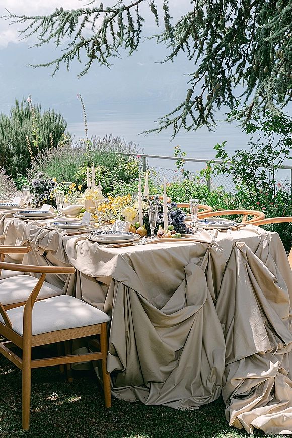 Reception tablescape with an outdoor wedding table setting of draped linen, taper candles, floral fruit centerpiece, and place cards by a lake and mountains