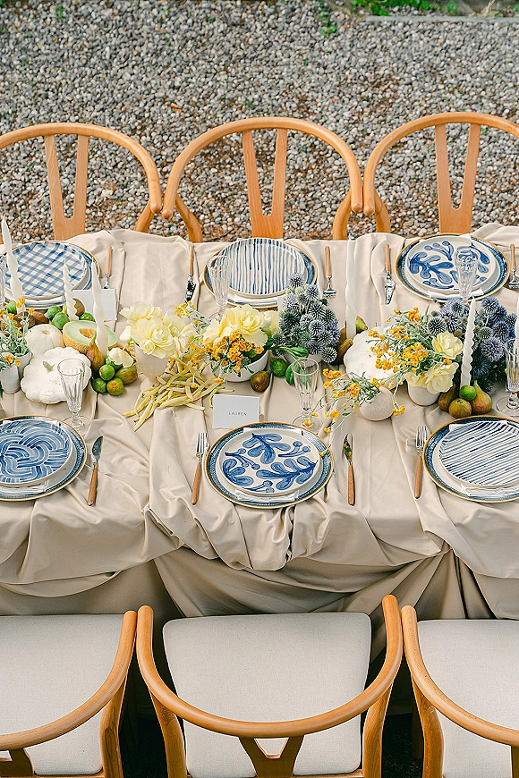 Reception tablescape with blue and white wedding plates on beige linen, crystal stemware, twisted taper candles, and seasonal produce accents outdoors on gravel