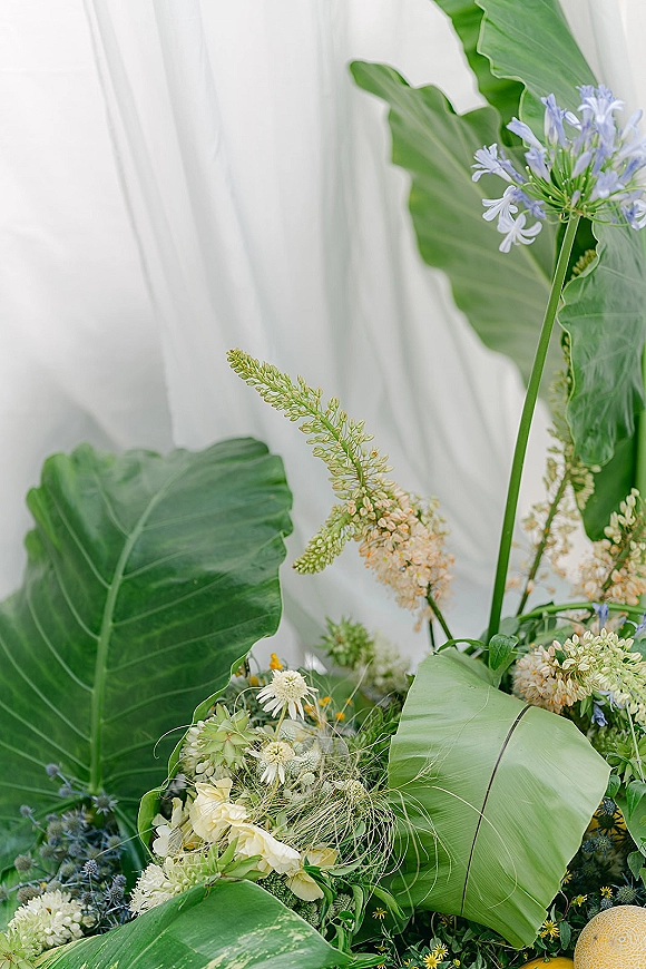 Wedding floral arrangement with tropical wedding flowers, banana leaves, white blooms, and blue accents against a draped white curtain backdrop