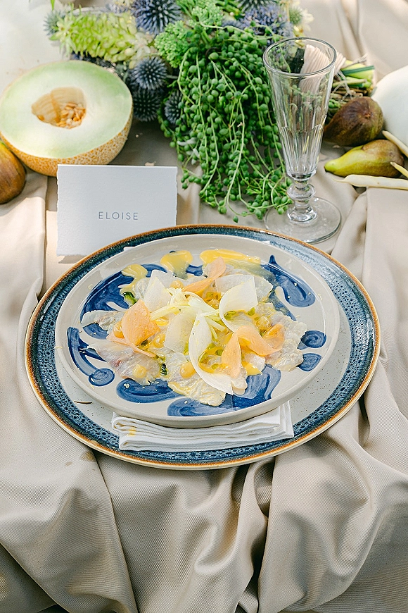Reception tablescape with wedding place setting, blue and white ceramic plates, folded napkin, place card, clear goblet, fruit and greenery accents