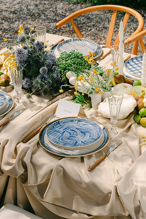 Reception tablescape with blue and white tablescape plates, cream linen, twist candles, thistle florals, fruit, and white pumpkins on gravel outdoors