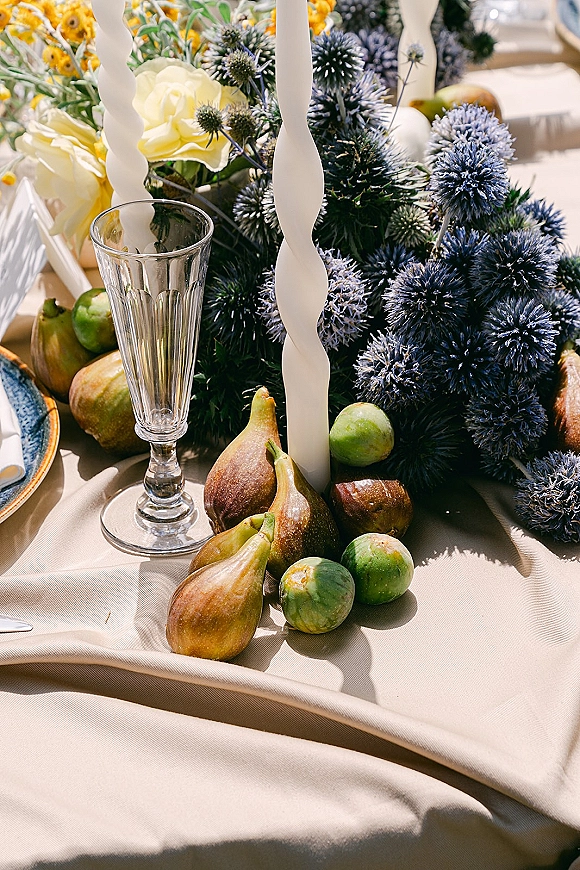 Reception tablescape with a wedding centerpiece with candles, thistle and yellow roses, figs, and a champagne flute in soft outdoor light