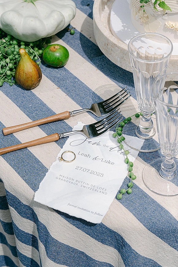Wedding invitation suite with a deckle edge wedding invitation, rings and fruit on a striped outdoor tablescape beside a small cake