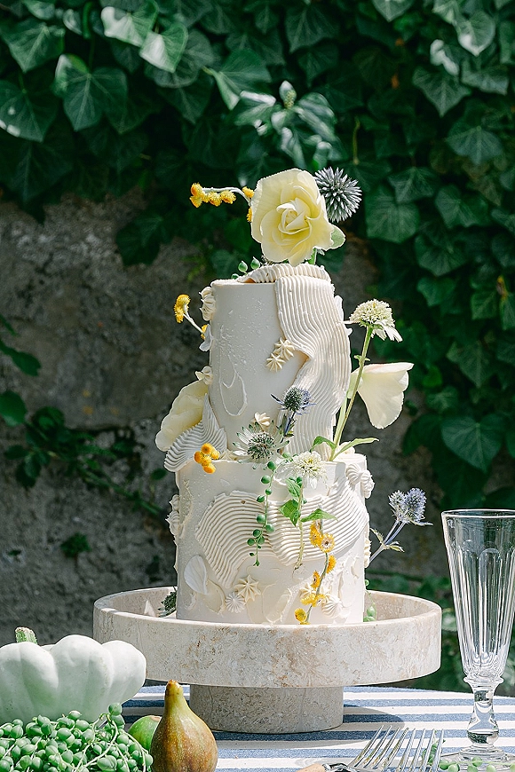 Wedding cake with textured buttercream on a stand, topped with fresh flowers and greenery, set against an ivy-covered stone wall outdoors