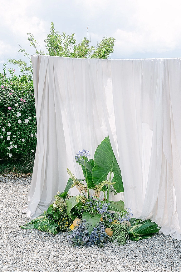 Ceremony backdrop with white draped wedding backdrop and greenery base florals, tropical leaves, and blue blooms on a gravel garden site