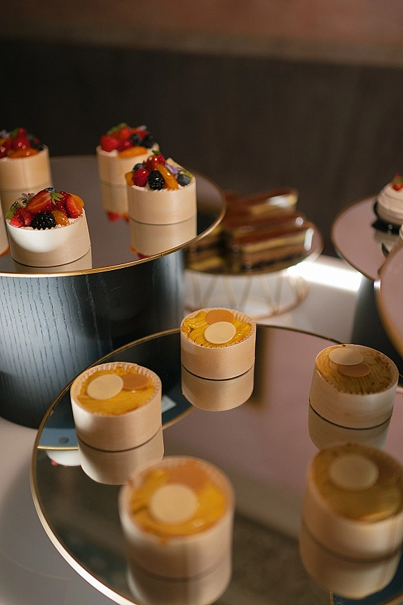 Wedding dessert table with mini wedding cakes topped with fruit on mirrored stands and gold rim trays in an elegant indoor reception space