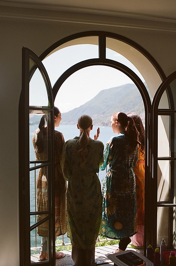 Bridesmaid getting ready in printed robe by a balcony railing, framed by open arched doors with lake and mountains beyond