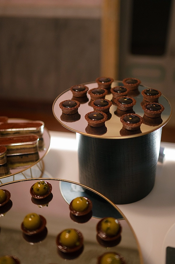 Wedding dessert table with mini dessert bar of bite-sized cups and chocolate tartlets on mirrored stands and gold-rim trays by a window