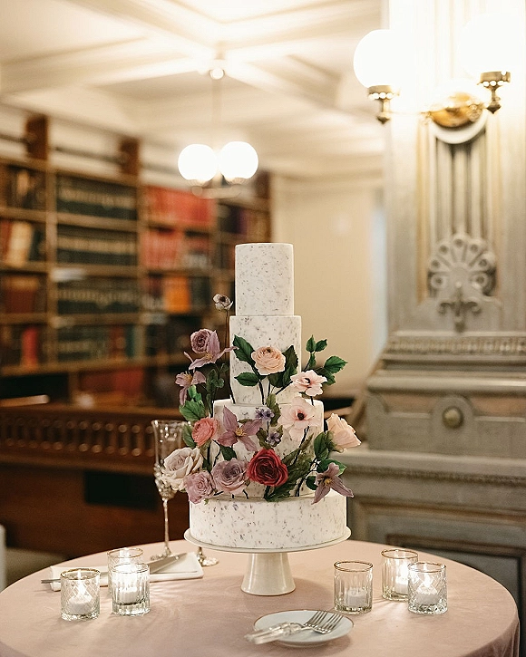 Wedding cake with tiered wedding cake buttercream frosting and rose florals on a cake stand, styled with candles by bookshelves and a fireplace mantel