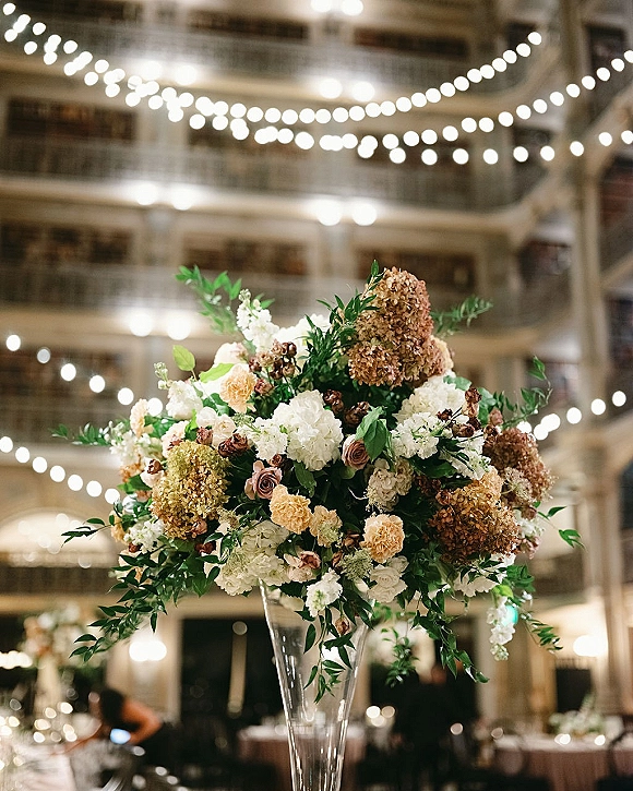 Wedding centerpiece with tall wedding centerpiece hydrangeas and roses in a glass vase under string lights in a softly lit reception hall