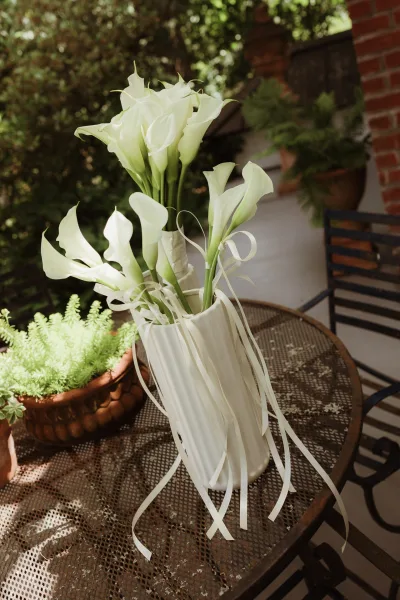 Calla lily bouquet of white calla lilies arranged in a ceramic vase on a metal table, framed by potted plants and a brick-walled garden patio.