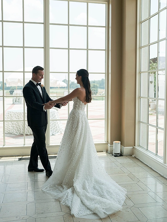 First look moment as bride in a strapless lace gown with long train holds groom’s hands by large windows overlooking water view