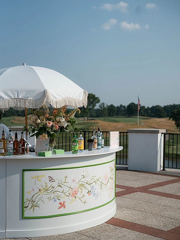 Outdoor wedding bar with a white fringe umbrella, floral arrangement, and liquor bottles on a round counter on a sunny patio terrace