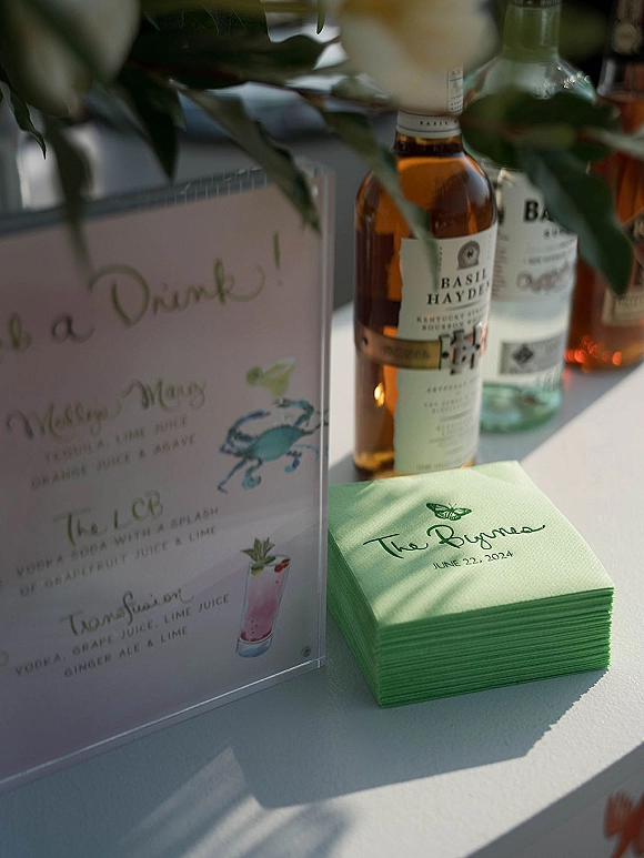 Signature drink sign with calligraphy wedding bar sign in a clear acrylic holder, framed by liquor bottles, napkins, and greenery on a sunlit bar table