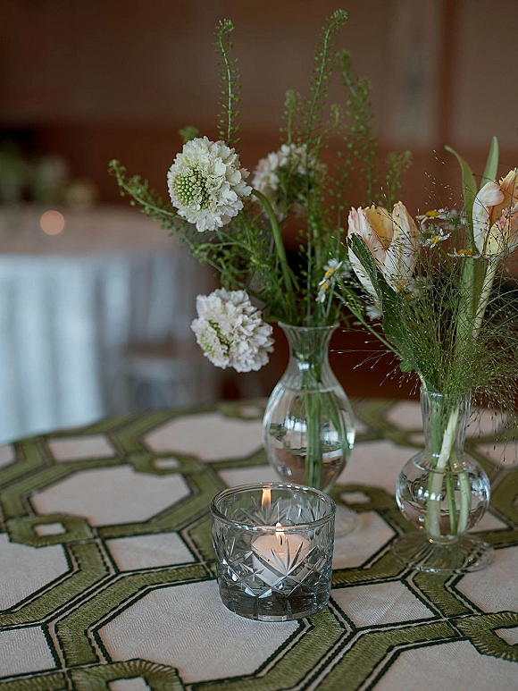 Reception tablescape with bud vase centerpieces, mixed wildflowers and greenery beside a glowing votive candle on patterned tablecloth indoors