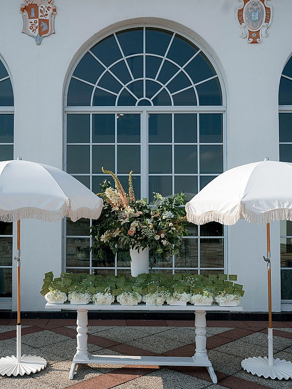 Escort card display on a white table with green escort cards and a large white vase floral arrangement, set by an arched window facade outdoors