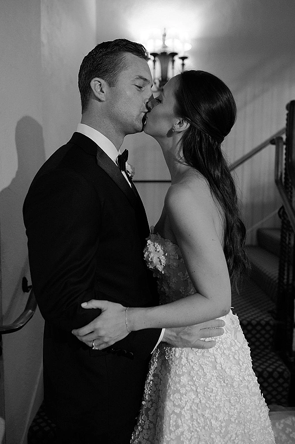 Wedding kiss portrait in black and white of bride in a strapless lace gown and groom in tuxedo on a staircase in a hallway