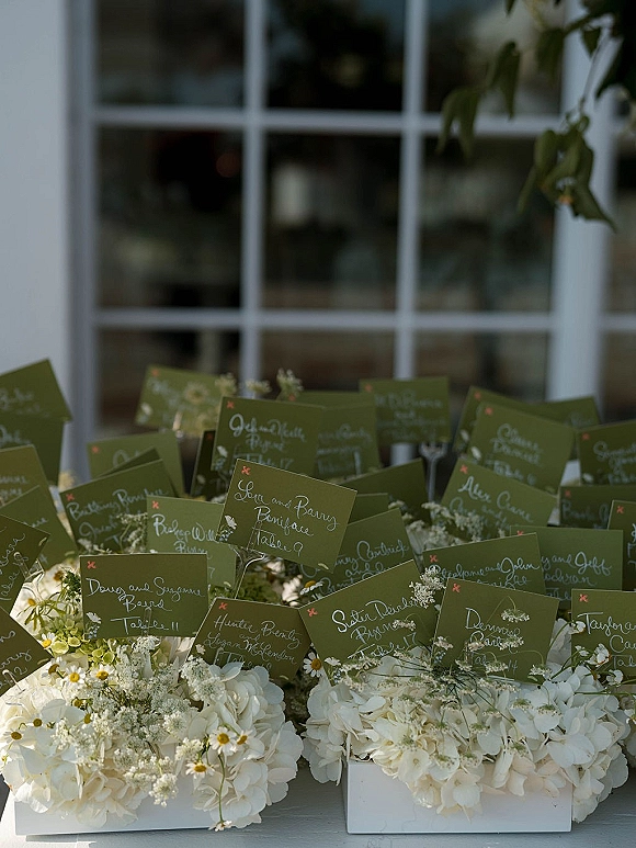Wedding escort cards in a lush escort card display with olive green place cards nestled among white hydrangeas by window panes